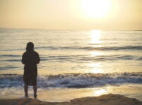 夕暮れの海辺に立つ人影と、波に反射する柔らかな光の情景｜懐かしい情景が静かによみがえる、やさしい記憶のイメージ写真