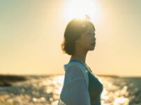夕暮れの海辺で、柔らかな逆光に照らされながら横顔で遠くを見つめる女性の姿｜懐かしい情景が静かによみがえる、やさしい記憶のイメージ写真