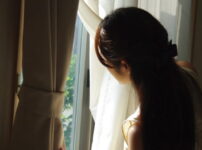 窓辺でやさしい光を受けながら外を見つめる女性の後ろ姿｜懐かしい情景が静かによみがえる、やさしい記憶のイメージ写真
